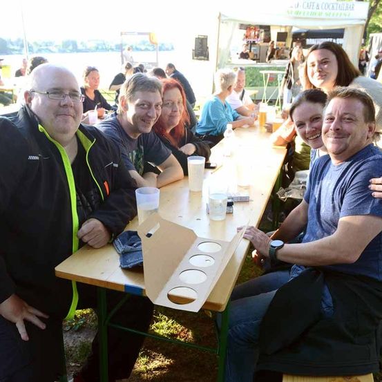 Eine Gruppe von Menschen sitzt im Freien um einen Tisch herum, lächelt und posiert für ein Foto. Sie haben Getränke und Essen auf dem Tisch. Hinter ihnen steht ein Zelt mit einem Schild und einigen Leuten. In der Ferne befindet sich ein Gewässer.