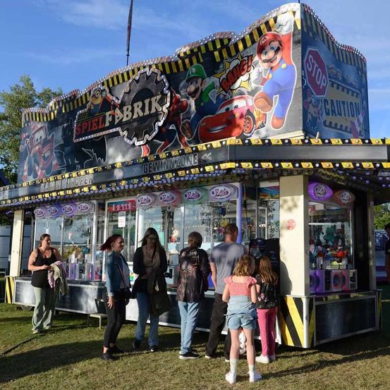 Eine Gruppe von Menschen steht vor einem farbenfrohen Spielstand, mit bunten Figuren und einem Stoppschild auf dem Dach.