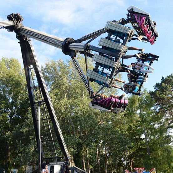 Eine große drehende Achterbahn mit mehreren Fahrgästen, die in der Luft aufgehängt sind, vor dem Hintergrund von Bäumen und blauem Himmel.