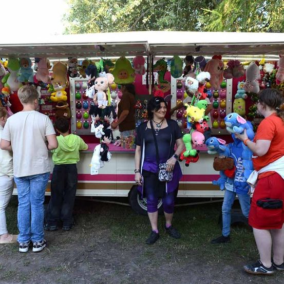 Eine Familie steht in einer Schlange, um ein Jahrmarktspiel zu spielen, bei dem sie Plüschtiere gewinnen können, während eine Frau hinter dem Tresen lächelt.
