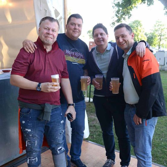 Vier Männer stehen draußen, lächeln und posieren für ein Foto. Einer hält ein Handy, ein anderer ein Bier. Im Hintergrund ist ein Zelt und Bäume zu sehen.