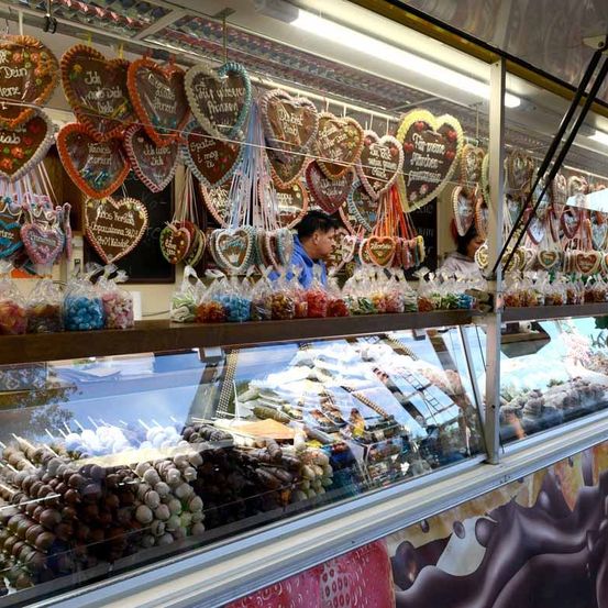 Eine Vitrine gefüllt mit verschiedenen Süßigkeiten, darunter herzförmige Kekse, Schokoladen und Lutscher. Zwei Personen stehen hinter der Theke, eine trägt ein blaues Hemd.