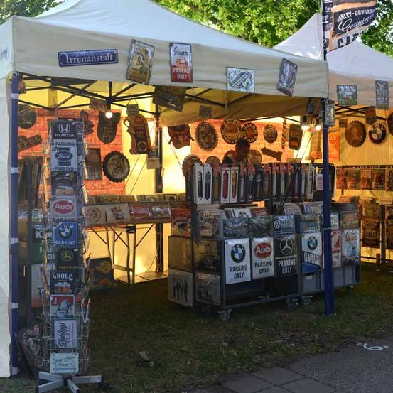 Ein Außenmarktstand zeigt Vintage-Schilder und Artikel unter einem weißen Zelt, mit einem Mann im Hintergrund. Der Stand präsentiert eine Vielzahl von Schildern und Produkten.