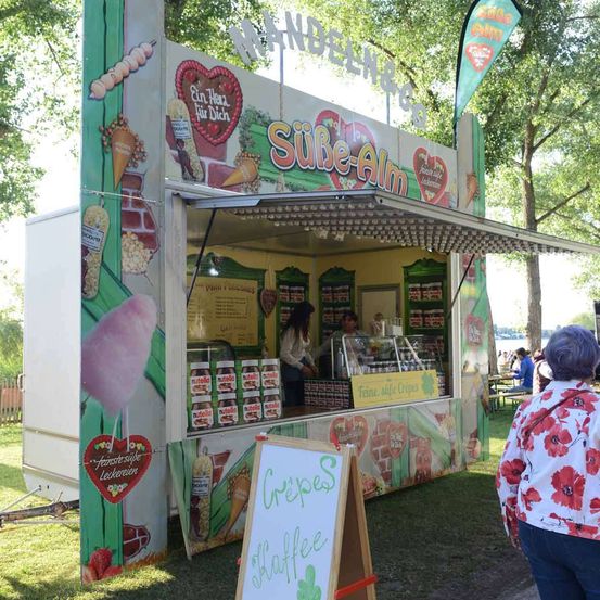 Ein Imbisswagen bietet süße Leckereien an. Eine Frau steht hinter der Theke, und ein Schild davor sagt Crepes und Waffee. Ein Kunde geht vorbei.