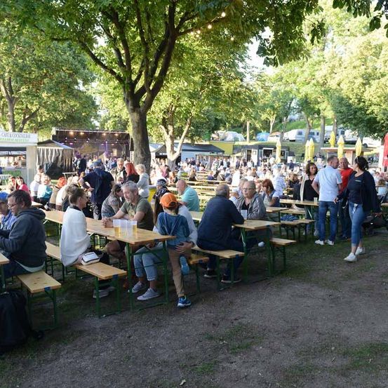 Eine große Menschenmenge versammelt sich in einem Park, sitzt an Bänken unter Bäumen. Einige haben Getränke in Gläsern. Ein Erwachsener trägt einen Hut, und ein Kinderwagen steht in der Nähe. Im Hintergrund gibt es Essensstände.