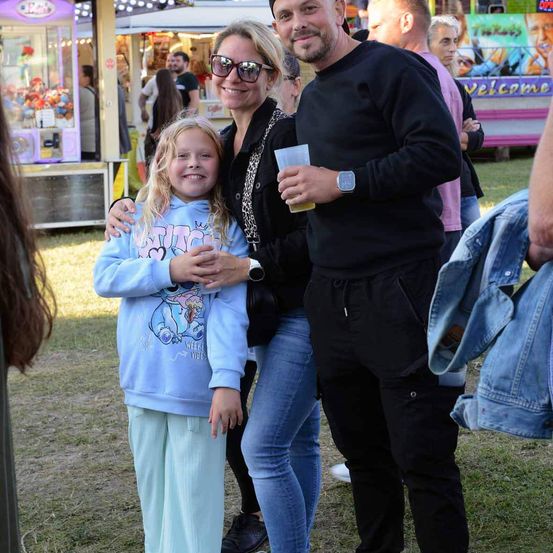 Ein Mann und eine Frau, mit einem lächelnden jungen Mädchen zwischen ihnen, posieren für ein Foto auf einem Jahrmarkt. Hinter ihnen befinden sich Karussells und eine Menge von Menschen.