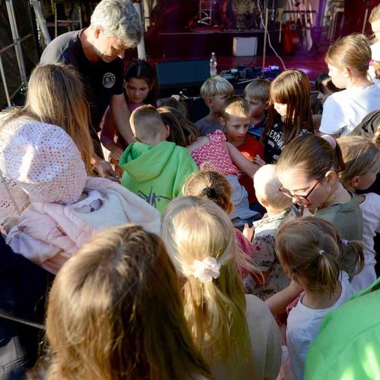 Eine Gruppe von Kindern und Erwachsenen versammelt sich um einen Mann, wahrscheinlich für eine Aufführung oder ein Ereignis, mit einer Bühne und Musikinstrumenten im Hintergrund.