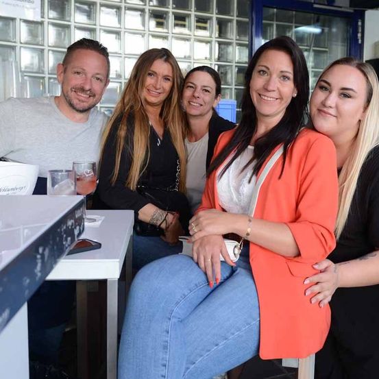 Fünf Menschen lächeln und posieren für ein Foto in einem Restaurant mit Glaswänden und einer blauen Tür. Ein Tisch mit Getränken und einer Schüssel steht vor ihnen.