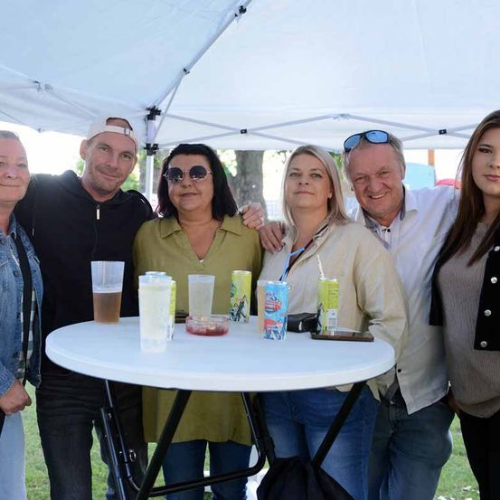 Eine Gruppe von Menschen steht unter einem weißen Zelt mit einem Tisch davor. Sie lächeln und posieren für ein Foto. Der Tisch hat Becher, Flaschen und andere Gegenstände darauf. Der Mann links trägt einen Hut und die Frau rechts einen Halsschmuck. Hinter ihnen sind Bäume und ein grasbewachsenes Feld.