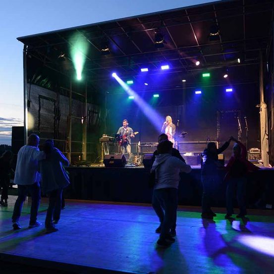 Eine Live-Performance auf einer Bühne mit bunten Lichtern, eine Frau singt und ein Mann spielt Gitarre. Mehrere Menschen tanzen vor der Bühne.