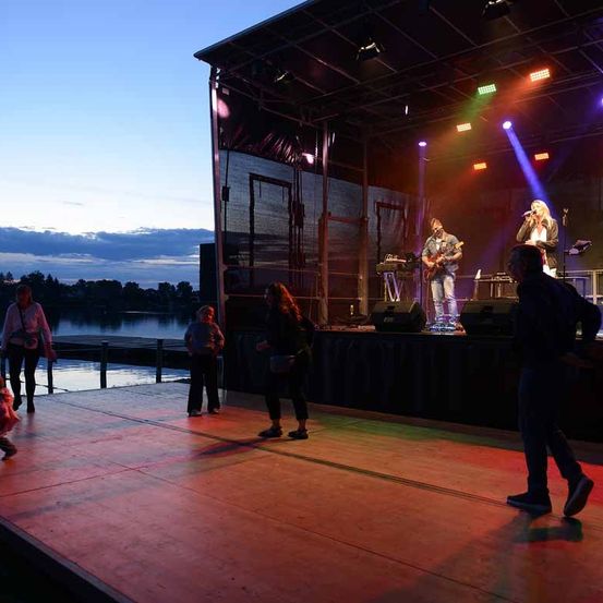 Eine Gruppe von Menschen tritt auf einer Bühne vor einem See auf. Die Bühne ist mit bunten Lichtern beleuchtet, und zwei Personen spielen Gitarre und singen. Das Publikum steht vor der Bühne.