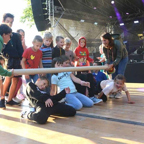 Eine Gruppe von Kindern versammelt sich auf der Bühne, einige knien und andere stehen. Eine Frau hält eine Trommel und ein langes Objekt. Die Bühne hat einen Holzboden und eine Kulisse mit lila Lichtern.