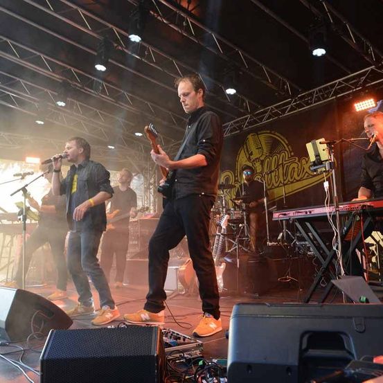 Eine Band spielt auf der Bühne, wobei ein Gitarrist die Hauptrolle spielt. Ein Sänger singt in ein Mikrofon. Dahinter spielt ein anderer Bandmitglied die Gitarre. Es gibt Lautsprecher, ein Schlagzeug und ein Keyboard auf der Bühne.