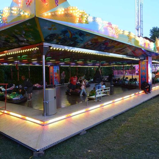 Eine beleuchtete Achterbahn mit buntem Dach ist bei Nacht. Einige Leute sitzen auf den Autos, während andere an der Seite stehen.