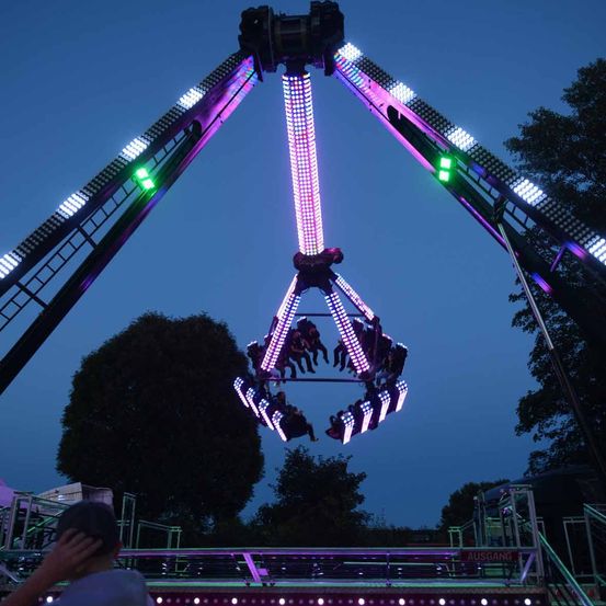 Eine Karussellfahrt ist nachts mit lila und grünen Lichtern beleuchtet. Eine Person beobachtet vom Boden aus.