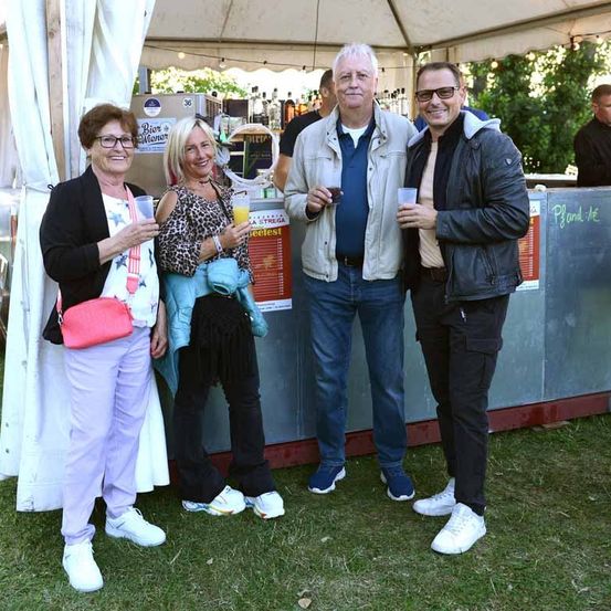 Vier Personen stehen unter einem Zelt, lächeln und halten Gläser in den Händen. Sie scheinen sich bei einer Veranstaltung im Freien zu befinden. Hinter ihnen ist eine Theke mit einem Menübrett zu sehen.