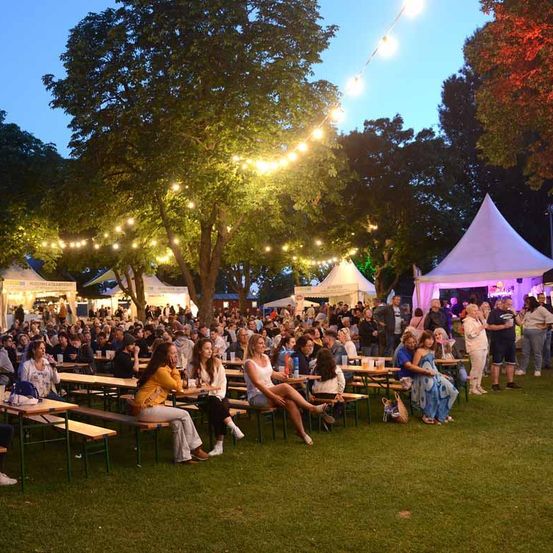 Bei Dämmerung versammelt sich eine große Menschenmenge im Freien unter Lichterketten und genießt Tische und Zelte. Einige sitzen, andere stehen, und Musik spielt im Hintergrund.