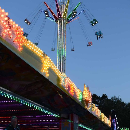 Beleuchtete Karussellattraktion mit bunten Lichtern, Menschen schwingen in der Luft, eine Person steht in der Nähe.