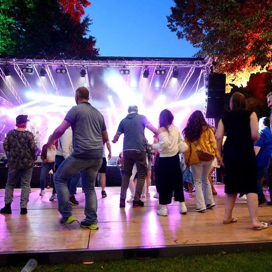 Eine Gruppe von Menschen tanzt auf einer Bühne mit Beleuchtung, wahrscheinlich bei einem Konzert, unter einem Himmel mit Bäumen.