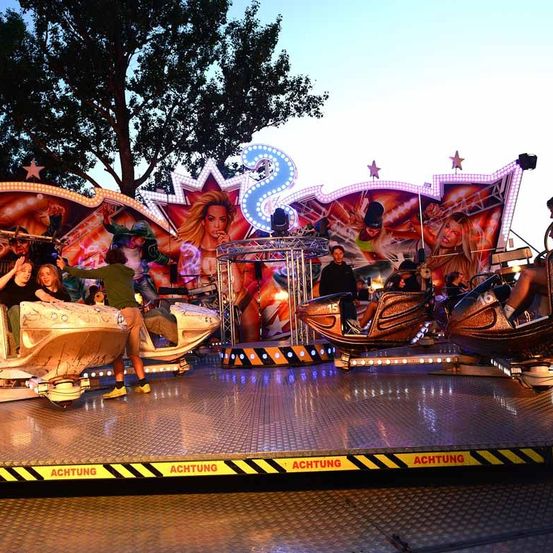 Eine Gruppe von Menschen genießt eine Fahrt auf einer farbenfrohen, neonbeleuchteten Jahrmarkt-Attraktion mit zwei Frauen, die auf dem Hintergrund der Attraktion abgebildet sind.
