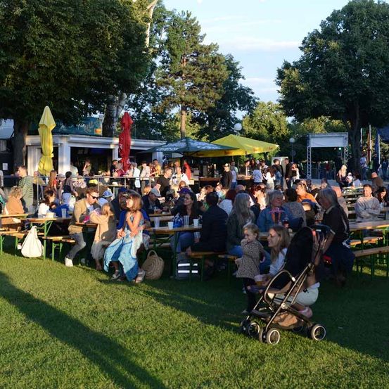 Eine große Gruppe von Menschen, einschließlich Familien, ist im Freien versammelt. Sie sitzen an Tischen unter Sonnenschirmen und Zelten und genießen Essen und Getränke. Bäume und eine Bühne sind im Hintergrund zu sehen.
