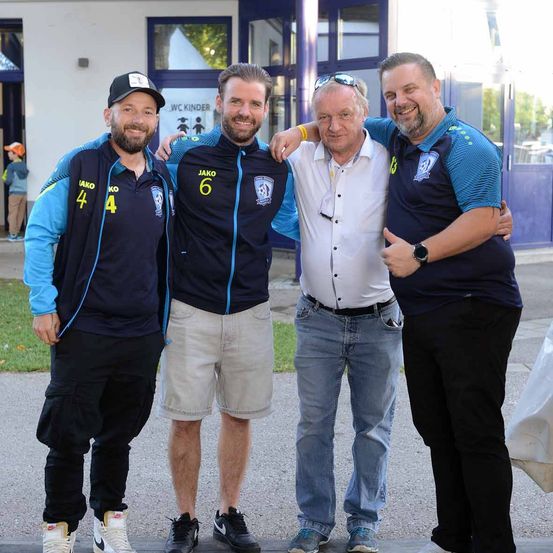 Vier Männer stehen auf einem Bürgersteig, drei tragen Trikots mit den Nummern 4, 6 und 3, wobei der Mann in der Mitte eine Sonnenbrille auf dem Kopf trägt. Sie scheinen für ein Foto zu posieren.