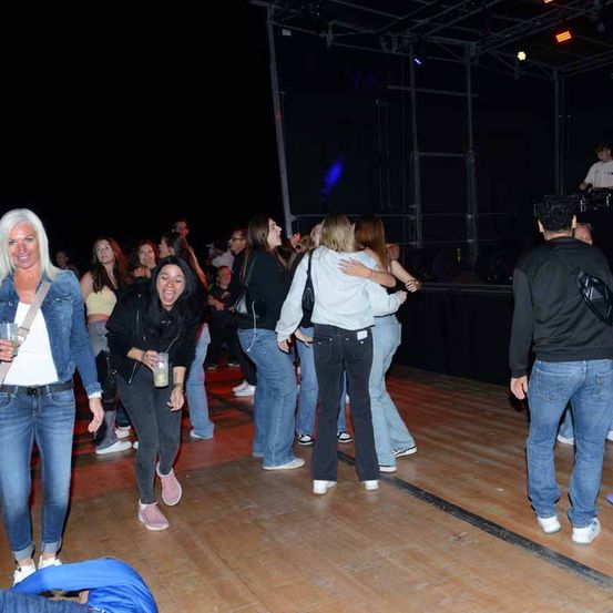 Eine Gruppe von Menschen tanzt in einem Club, einige tragen Turnschuhe. Eine Frau hält ein Getränk. Eine Person in einem weißen Shirt steht hinter einem DJ-Set.