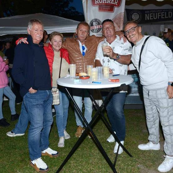 Fünf Personen stehen draußen auf einer Rasenfläche mit einem weißen Tisch. Sie lächeln alle und posieren für ein Foto. Auf dem Tisch stehen Getränke und Essen.