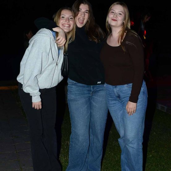 Drei junge Frauen stehen nachts auf einem Rasen und posieren für ein Foto. Die Frau auf der linken Seite trägt eine weiße Kapuzenjacke und schwarze Hose. Die Frau in der Mitte trägt ein schwarzes Oberteil und Jeans. Die Frau auf der rechten Seite trägt ein braunes Oberteil und blaue Jeans.