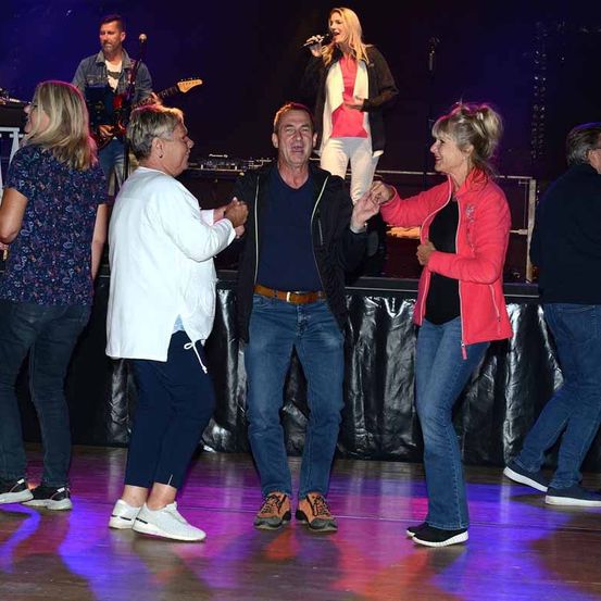 Eine Band spielt auf der Bühne mit einem Sänger. Ein Mann und zwei Frauen tanzen vor der Bühne, während ein anderer Mann und eine Frau zusehen.