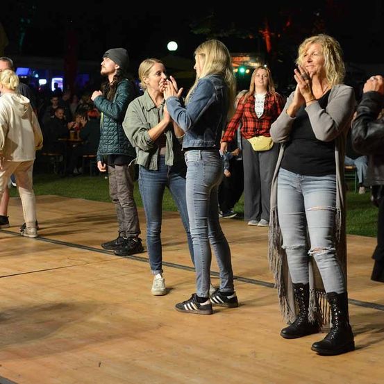Mehrere Frauen und Männer stehen nachts auf einer Holzbühne, klatschen und lächeln. Im Hintergrund ist eine verschwommene Menge und Tische mit Getränken zu sehen.