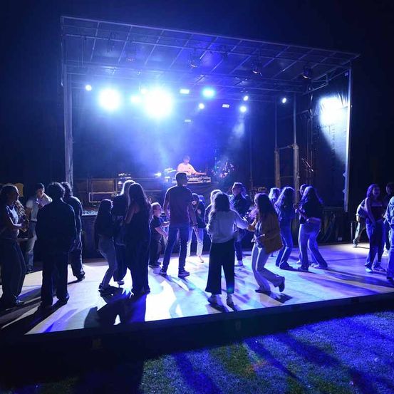 Eine nächtliche Aufführung mit einer von blauen Lichtern erleuchteten Bühne. Ein Mann spielt ein Instrument, während Leute um ihn herum tanzen.