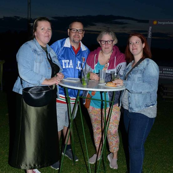 Vier Erwachsene stehen um einen kleinen Tisch herum, lächeln und posieren für ein Foto. Eine Person trägt eine blaue Jacke mit 'ITA' darauf. Getränke und Essen sind auf dem Tisch.