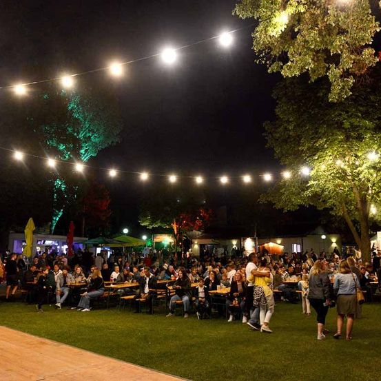 Ein nächtliches Open-Air-Festival mit Lichterketten und Bäumen. Viele Menschen sitzen auf Bänken und Tischen, während andere gehen.