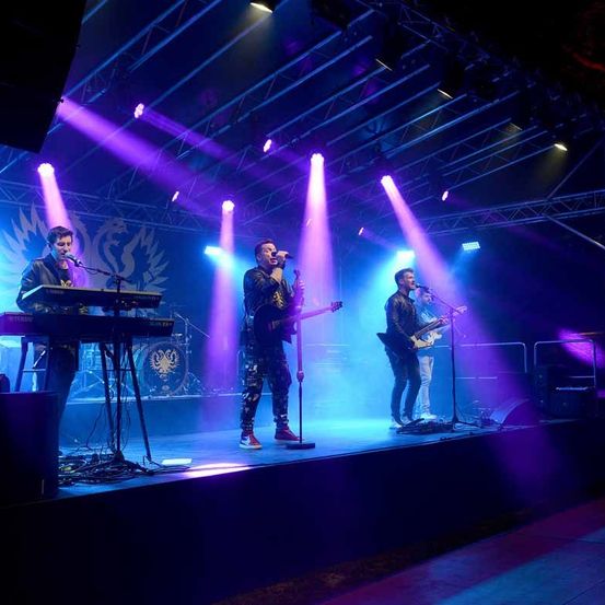 Eine Band tritt auf der Bühne unter lila und blauen Lichtern auf. Ein Mitglied singt, während ein anderes die Gitarre spielt. Ein weiteres Mitglied spielt Klavier.