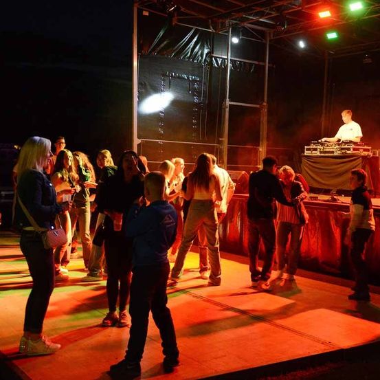 Eine Nachtveranstaltung mit einem DJ im Rampenlicht und Leuten, die auf einer Bühne mit roter Beleuchtung und Lautsprechern im Hintergrund tanzen.