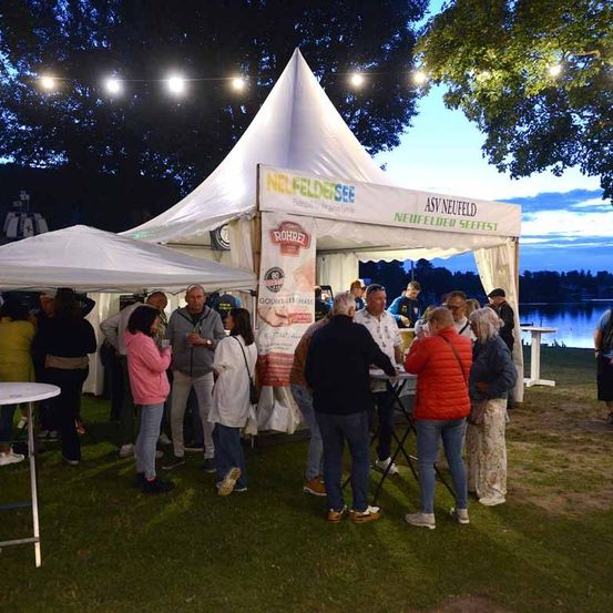 Menschen versammeln sich um ein weißes Zelt mit dem Wort Neufelder See. Sie sind in der Nähe eines Sees mit Bäumen und Lichtern im Hintergrund.