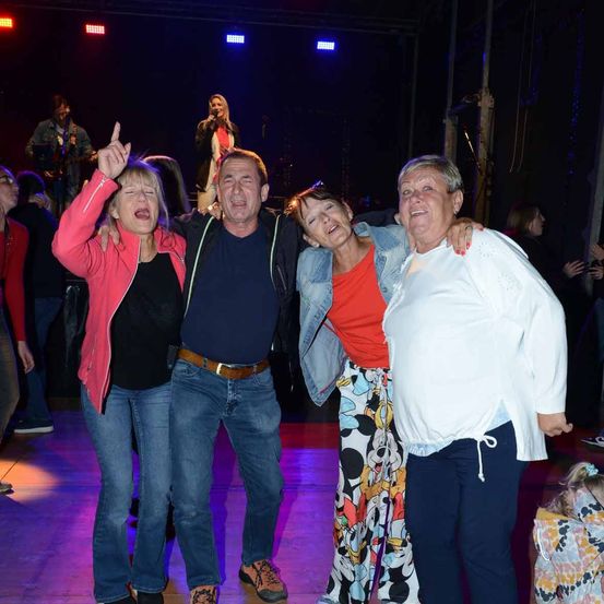 Vier Erwachsene posieren für ein Foto auf einer Bühne mit mehrfarbigen Lichtern. Eine Frau singt hinter ihnen, während andere zusehen.