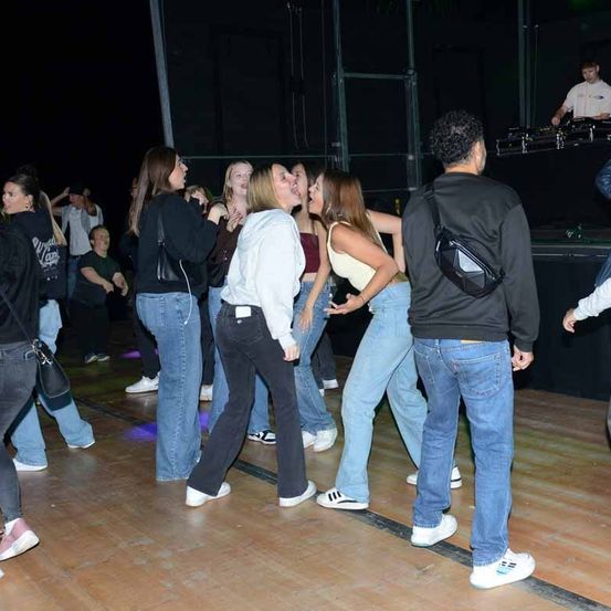 Eine Gruppe von Menschen tanzt in einem schwach beleuchteten Raum. Einige tragen Turnschuhe, andere Sandalen. Ein DJ bedient eine DJ-Anlage auf einer Bühne im Hintergrund.