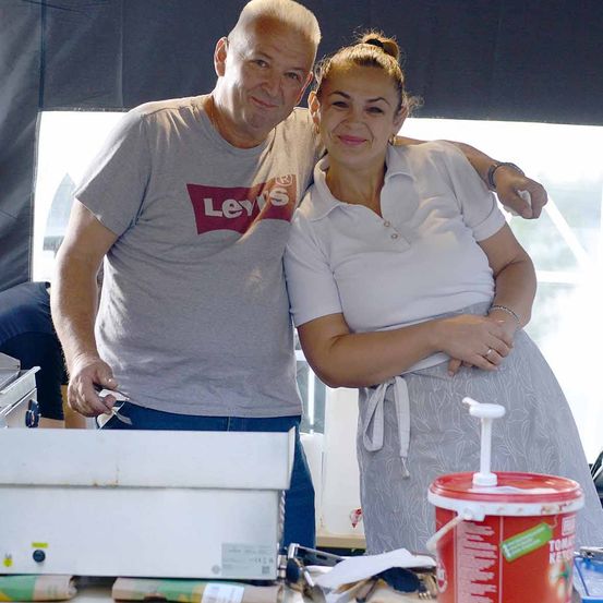Ein Mann und eine Frau lächeln für ein Foto bei einer Outdoor-Veranstaltung. Der Mann trägt ein Levi's-Shirt und blaue Jeans, während die Frau eine weiße Schürze trägt. Sie stehen neben einem Tisch mit einem roten Eimer.