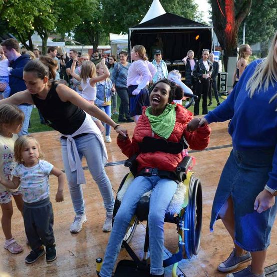 Eine Gruppe von Menschen tanzt im Freien. Eine Frau im Rollstuhl lächelt und lacht. Eine Frau in einem schwarzen Shirt hält ihre Hand. Mehrere Menschen stehen hinter ihnen, und es gibt Zelte und Bäume im Hintergrund.