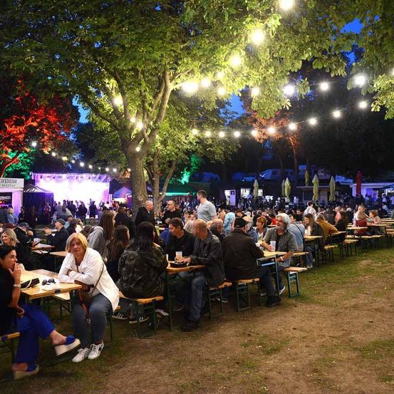 Ein Outdoor-Festival bei Nacht mit vielen Menschen, die an Tischen unter hängenden Lichtern sitzen, einer Bühne mit einer großen Leinwand und Bäumen und Sonnenschirmen drumherum.