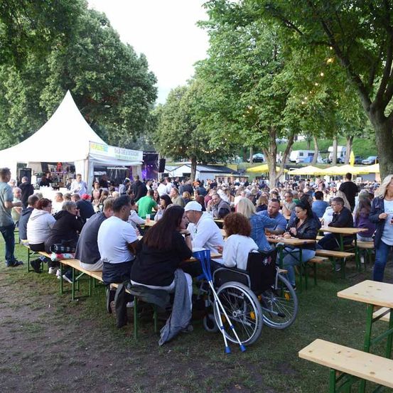 Eine große Menschenmenge versammelt sich in einem Park unter Zelten für eine Veranstaltung im Freien. Die Leute sitzen an Bänken und Tischen, einige in Rollstühlen, während andere in der Nähe von Essensständen und einer Bühne stehen.