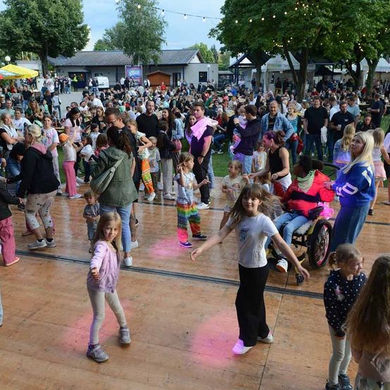 Eine große Menschenmenge ist auf einem Freigelände versammelt. Sie tanzen auf einem Holzboden. Einige halten ihre Kinder. Einige Bäume und Gebäude sind im Hintergrund.