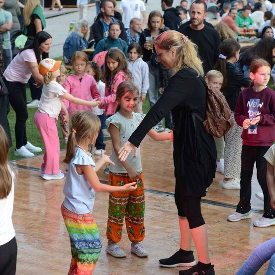 Eine Veranstaltung im Freien, bei der eine Frau Kinder zu einem Tanz führt, umgeben von einer großen Menschenmenge aus Erwachsenen und Kindern.