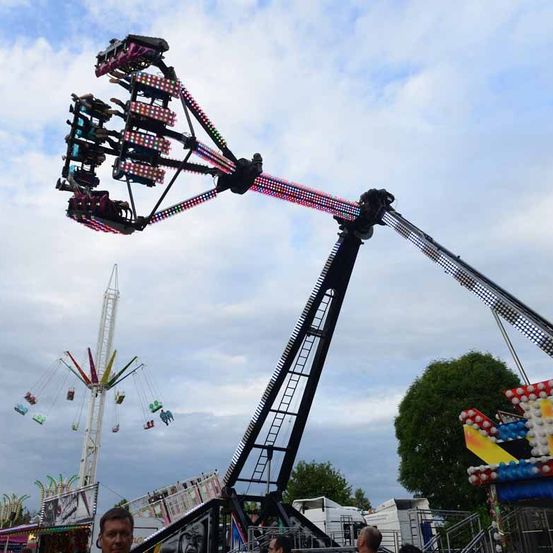 Eine große, farbenfrohe Achterbahn ist in der Luft aufgehängt und hat mehrere Sitze und leuchtende Lichter. In der Nähe sind andere Fahrgeschäfte und Attraktionen zu sehen, mit einem Baum und einem bewölkten Himmel im Hintergrund.
