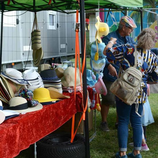 Ein mit rotem Samt bedeckter Tisch zeigt eine Vielzahl von Hüten, während ein erwachsenes Paar sich umsieht. Ein grünes Zelt ist darüber und ein Anhänger ist im Hintergrund zu sehen.