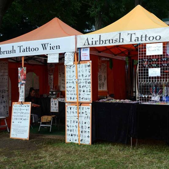 Zwei luftunterstützte Zelte für Airbrush-Tattoos sind in einem Grasbereich aufgestellt. Ein Zelt hat ein Banner mit der Aufschrift Airbrush Tattoo, während das andere Brush Tattoo Wien trägt. Eine Frau sitzt in einem Stuhl in einem der Zelte.