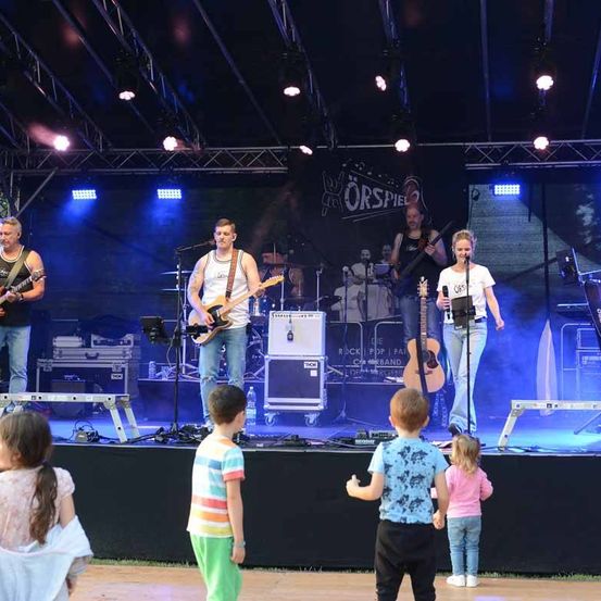 Eine Band spielt auf einer Bühne mit blauer Beleuchtung. Vier Musiker spielen Gitarre und Schlagzeug. Eine Menge Kinder und Erwachsene sieht ihnen zu.