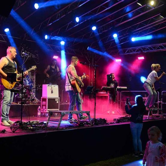 Eine Band tritt auf einer Bühne mit dem Hintergrund von Corsfield 2 auf. Sie werden von blauen und pinken Lichtern beleuchtet. Eine Frau macht ein Foto von der Band.